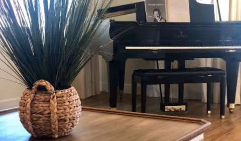 A black Kimball grand piano with a matching bench placed on a wooden floor. Next to the piano is a woven basket planter containing tall green grass-like plants. Light-colored curtains are visible in the background.