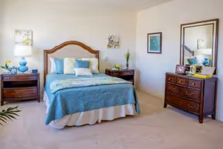 A neatly arranged bedroom featuring a bed with a wooden headboard, blue bedding, and multiple pillows. On either side of the bed are wooden nightstands with lamps and decorative items. A wooden dresser with a mirror stands against the wall, adorned with framed photos and small decorations. The room has light-colored walls and carpeted flooring.