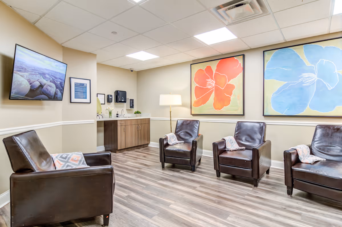 A cozy seating area with four dark brown leather armchairs, each with a patterned throw blanket draped over one arm. The room has light beige walls with white trim, two large colorful floral paintings on the wall, a flat-screen TV mounted on the left wall, and a wooden cabinet with a countertop and a lamp in the corner. The floor is covered with wood-patterned vinyl planks.