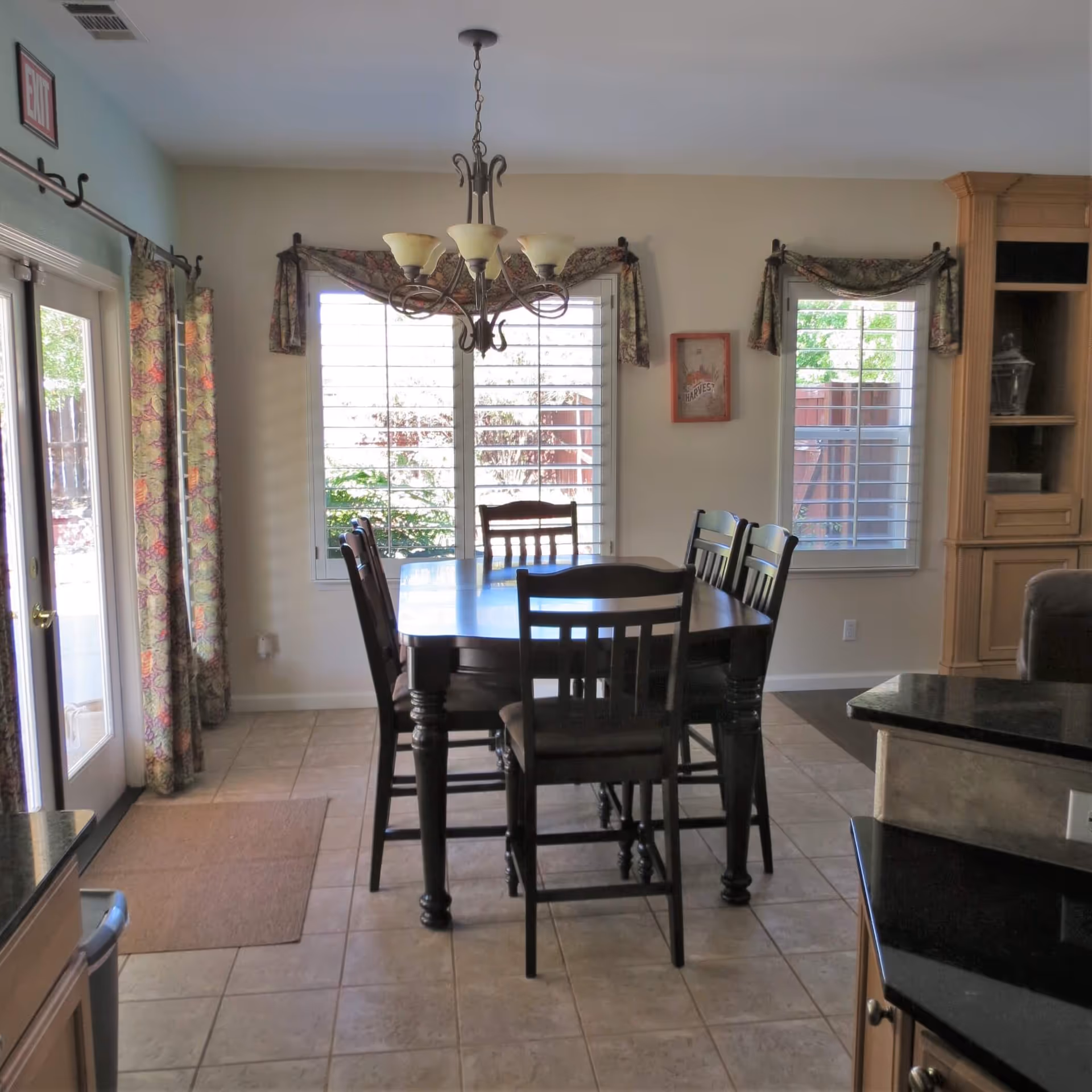 Dining area with a dark wooden table and six matching chairs on a tiled floor. The room has two large windows with white shutters and floral valances, a chandelier hanging above the table, and a glass door with floral curtains leading outside. There is a wooden cabinet on the right side and a kitchen counter with a black countertop partially visible.