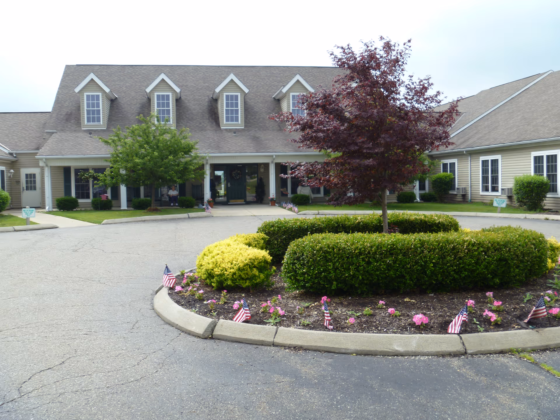 Front entrance of a two-story residential building with a circular driveway, landscaped island, and small American flags.