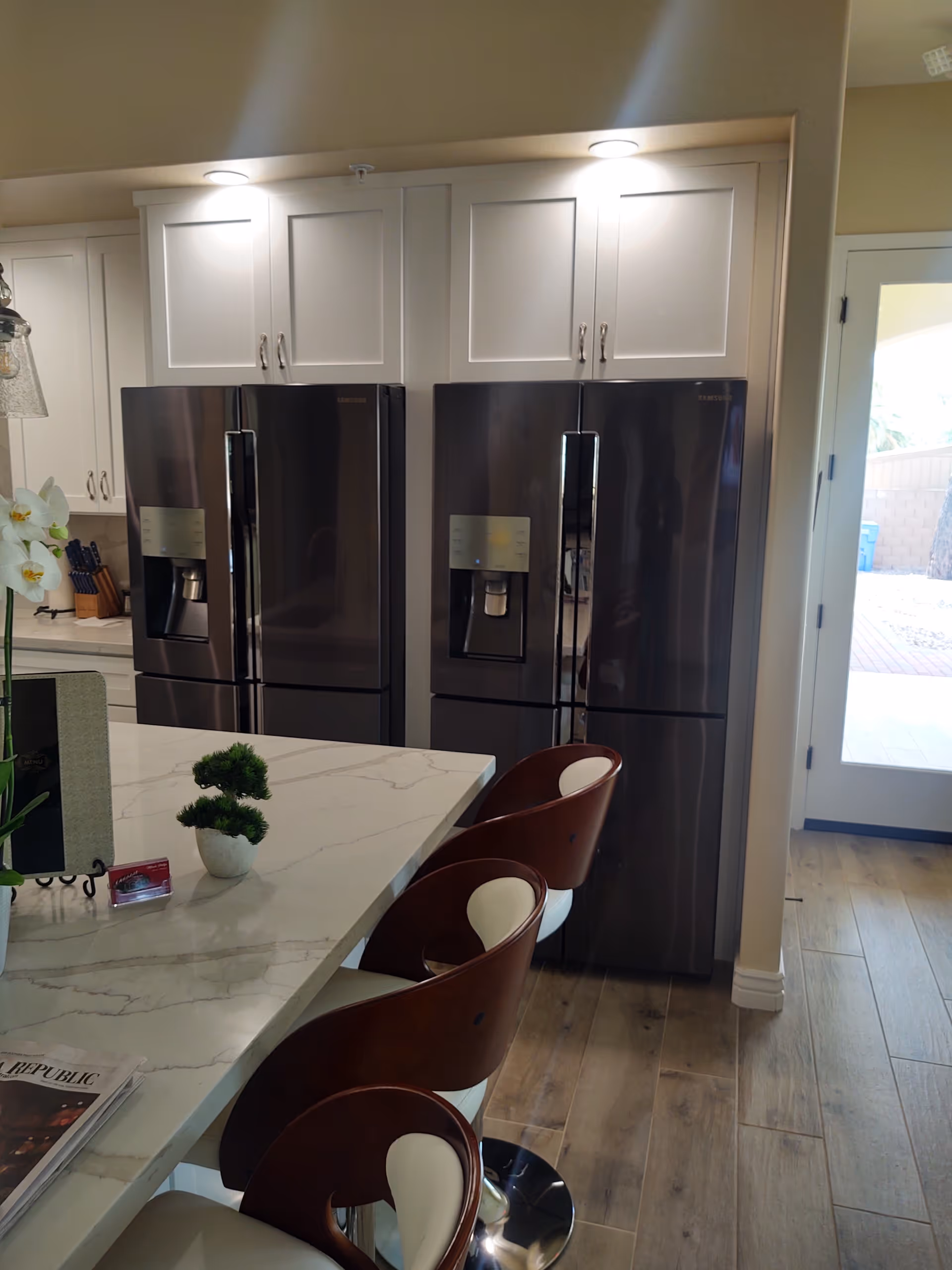 Modern kitchen area with two stainless steel refrigerators side by side, white cabinetry above them, a marble countertop island with three brown and white bar stools, a small potted plant, and a door leading outside.