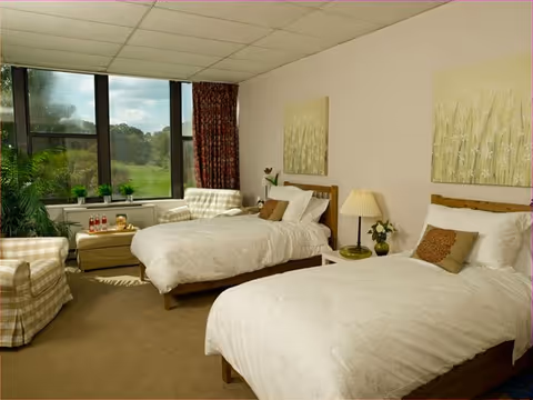 Bright twin bedroom with two single beds, bedside tables, armchairs and a large window overlooking greenery.