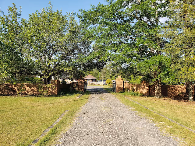 A gravel driveway flanked by green grass and large leafy trees leads to a gated entrance with brick pillars. Beyond the gate, there is a small building and more greenery under a clear blue sky.