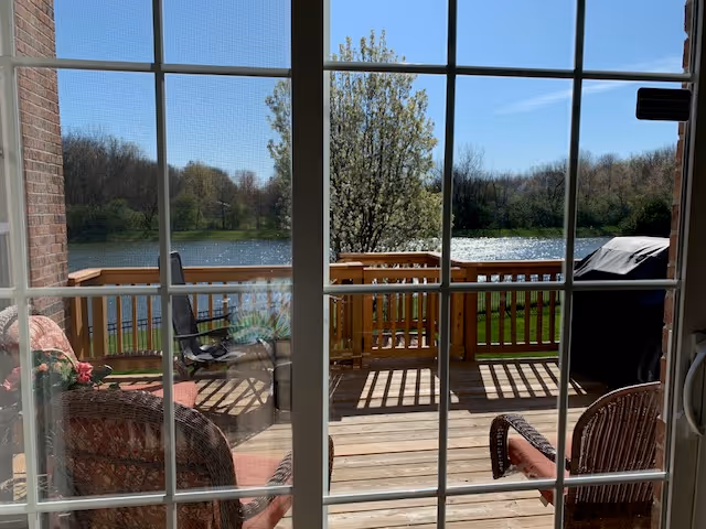 View through a sliding glass door onto a wooden deck with patio chairs and a grill overlooking a pond and trees.