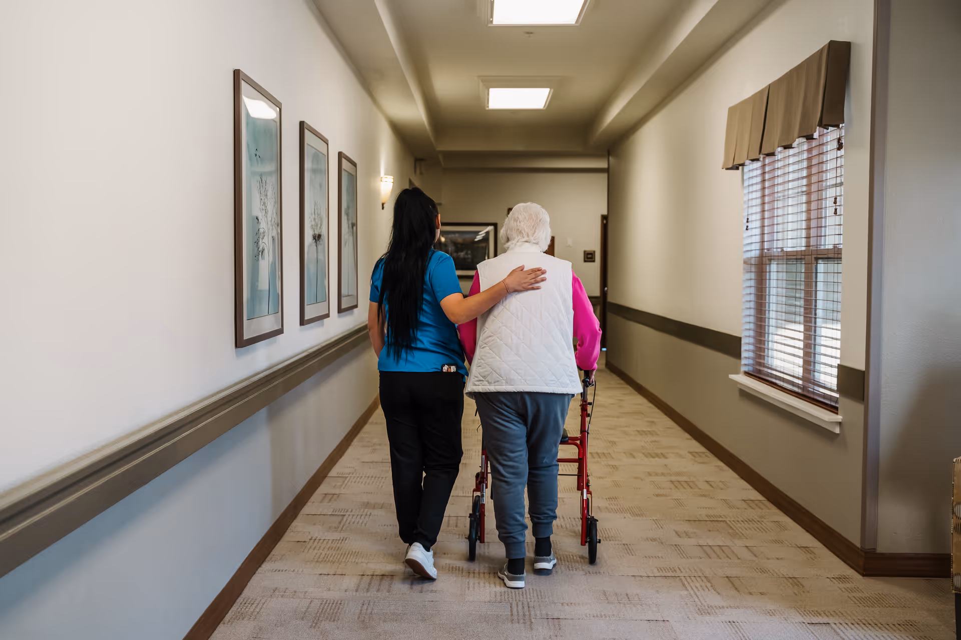 A caregiver walking down a hallway with an elderly woman using a walker. The caregiver has her arm around the woman for support. The hallway has framed pictures on one wall and a window with blinds on the other.