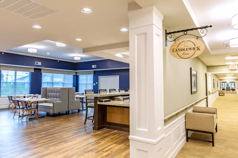 Interior view of a senior living facility dining area named Candlewick Bistro, featuring tables and chairs, a booth seating area, a granite countertop bar with stools, wood flooring, and a hallway with beige carpeting and chairs along the wall.