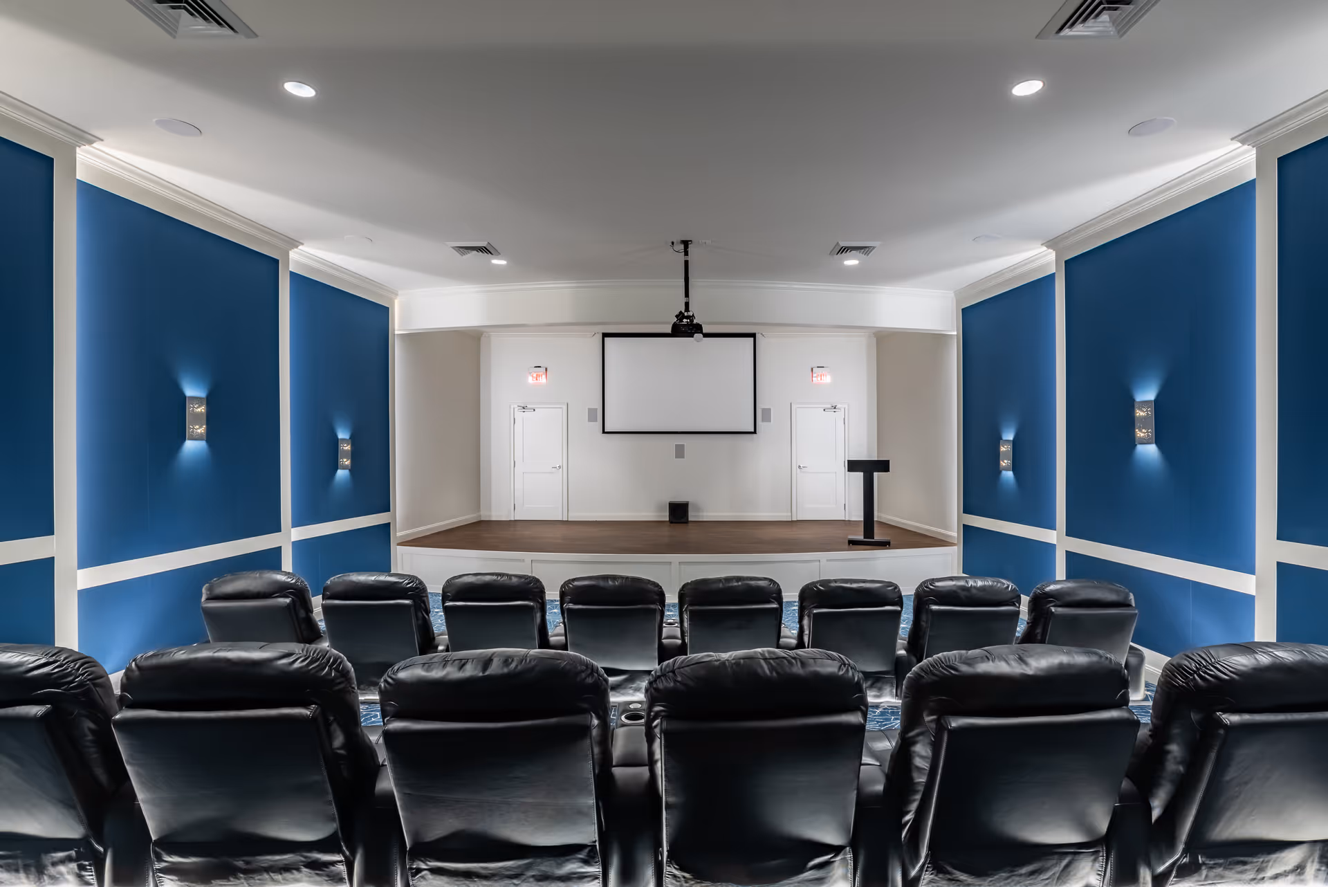 A small auditorium or screening room with two rows of black leather recliner chairs facing a stage with a projector screen and a podium. The walls are painted blue with white trim and have modern wall sconces providing light.