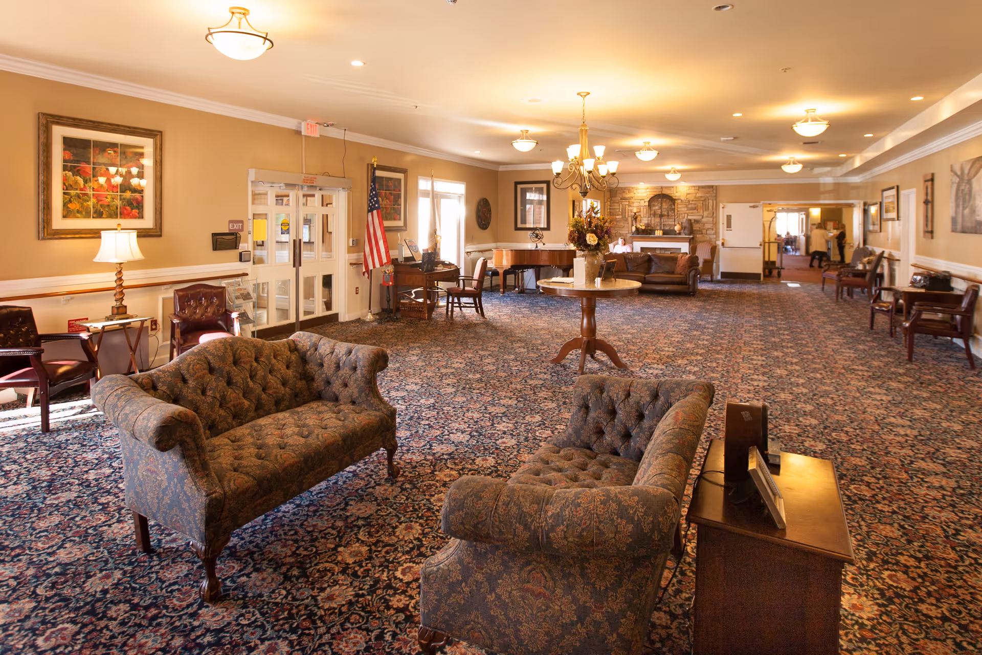 A spacious and warmly lit common area with patterned carpet, two upholstered sofas facing each other, a round table with a flower arrangement in the center, several chairs along the walls, framed artwork, an American flag near double glass doors, and a stone fireplace in the background.