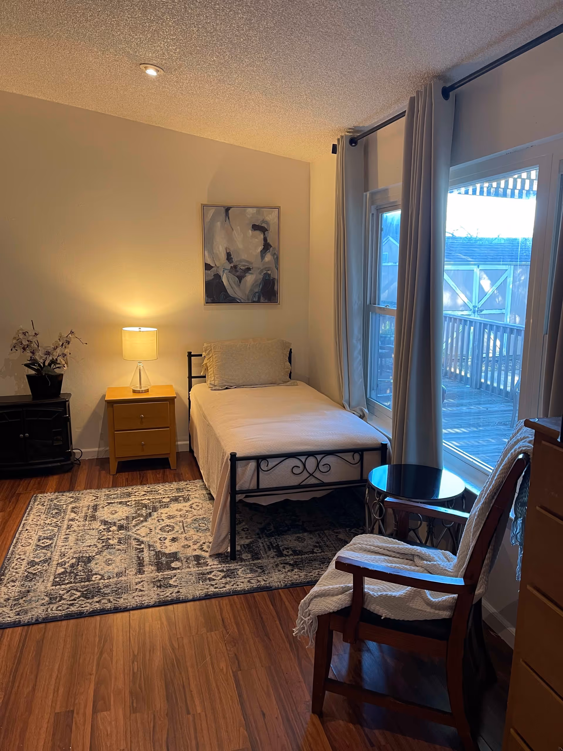 Cozy bedroom with a single metal bed, nightstand and lamp, a rug, chair and side table beside large windows overlooking a deck.