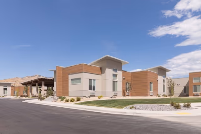 Low-rise modern senior living building with wood and light-colored siding, landscaped lawn, and a clear blue sky.