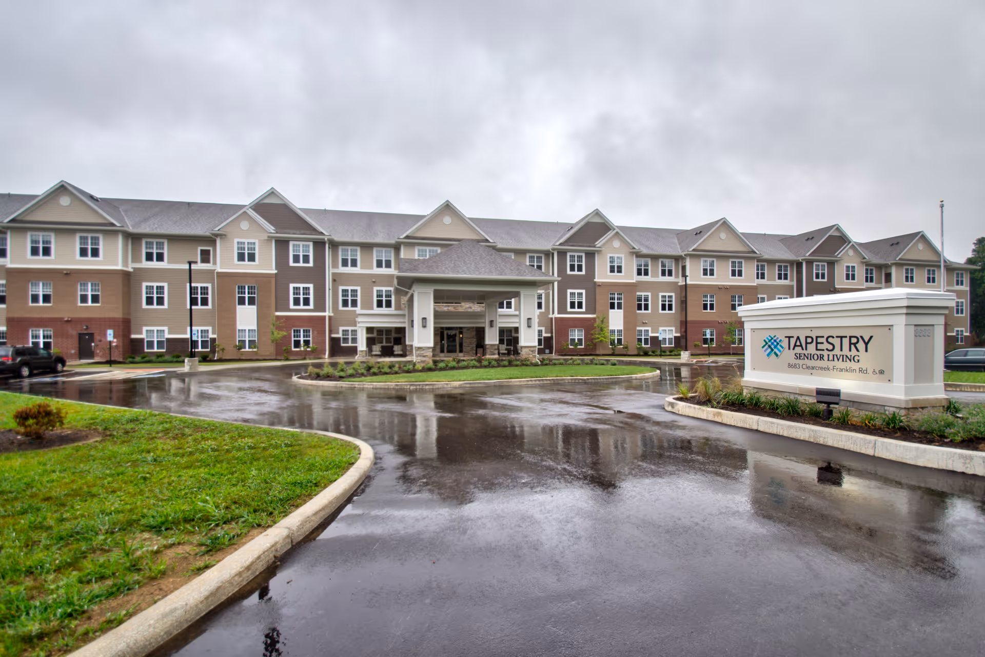 Front exterior of a three-story senior living building with a circular driveway and a Tapestry Senior Living sign on a rainy day.