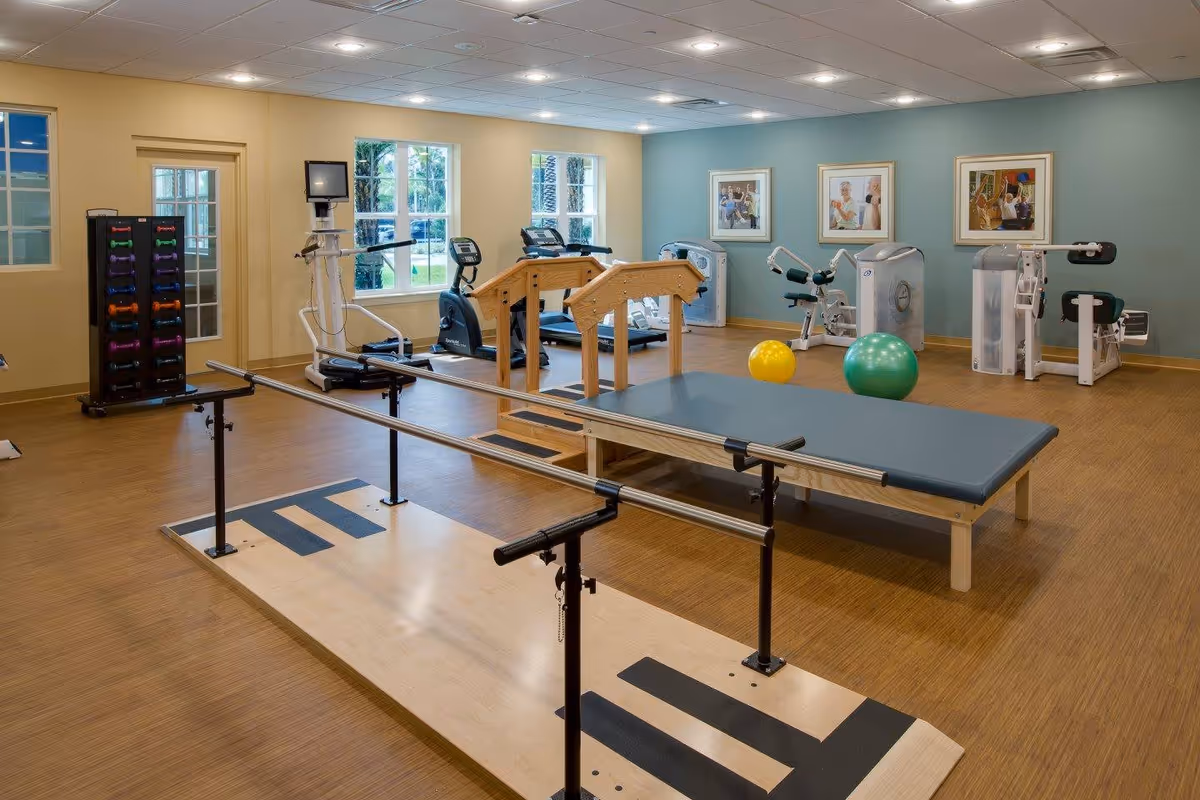 A spacious fitness and rehabilitation room with wooden flooring, parallel bars for walking exercises, a padded therapy table, exercise machines, colorful exercise balls, and a rack of dumbbells. The room has large windows letting in natural light and framed artwork on the walls.