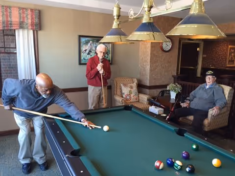 Three elderly men in a common room playing pool. One man is taking a shot with a pool cue, another man is standing and holding a cue stick, and the third man is sitting in a chair watching. The room has a pool table, hanging lights above it, a window with curtains, and comfortable seating.