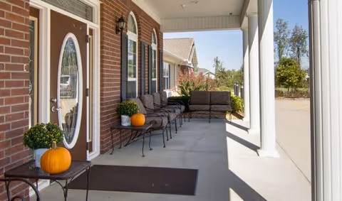 Covered outdoor patio area at Foxberry Terrace Senior Living with brick walls, a brown door with an oval glass window, black metal chairs with cushions, small tables holding pumpkins and potted plants, and white columns supporting the roof. Trees and a parking lot are visible in the background.