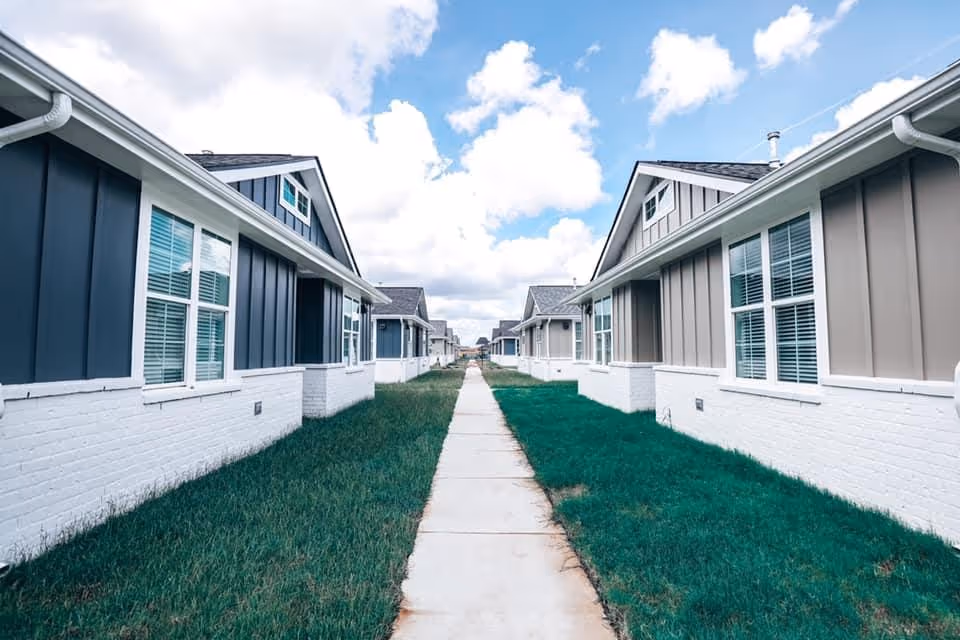 A central sidewalk runs between rows of single-story residential buildings with white brick bases and blue-gray siding under a partly cloudy sky.