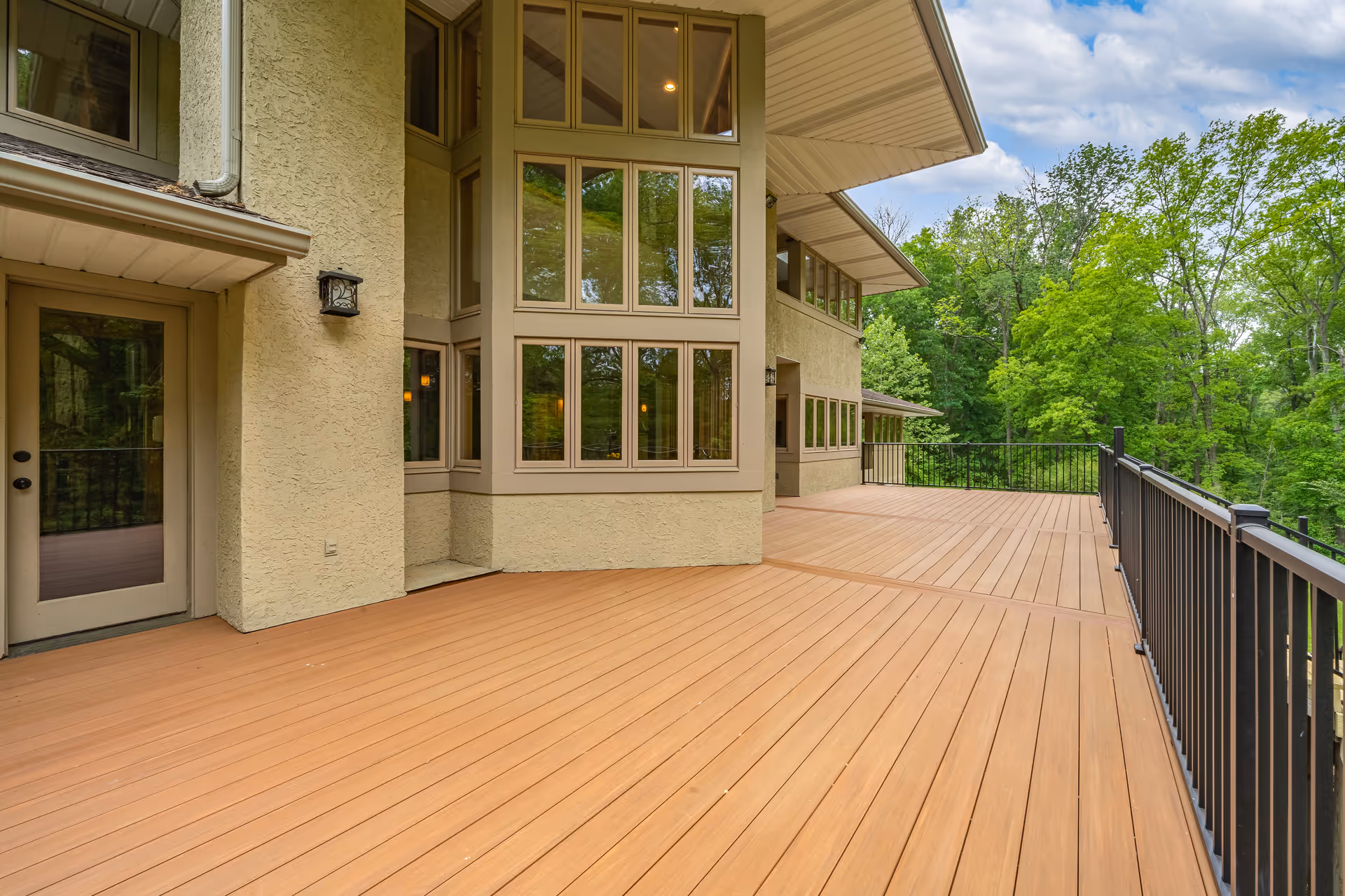 Spacious outdoor deck area with wooden flooring and black metal railing attached to a beige stucco building with large windows, surrounded by green trees under a partly cloudy sky.