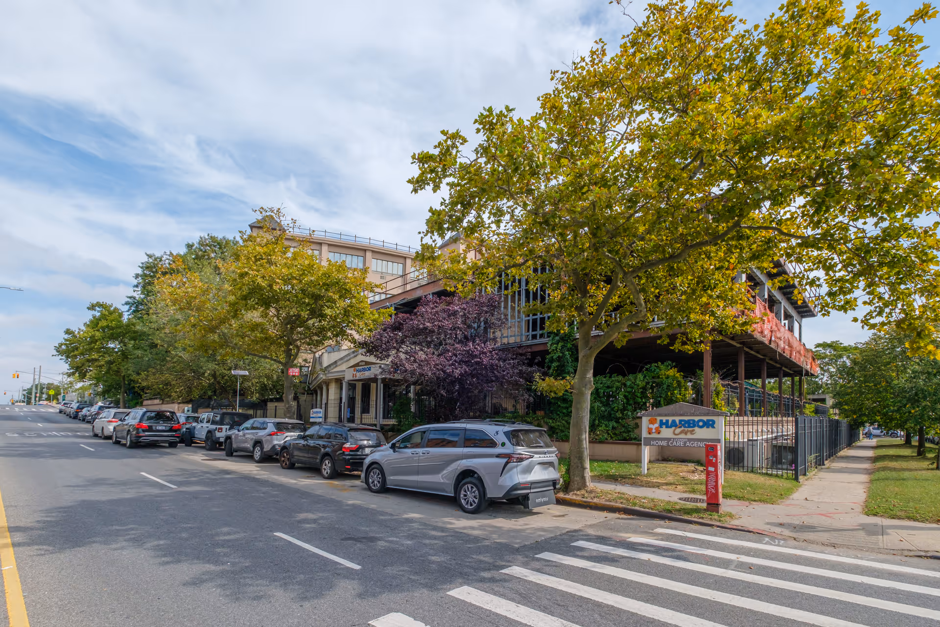 Street view of Harbor View Home, a multi-story building partially obscured by trees with green and purple foliage. Several cars are parked along the street in front of the building. A sign near the sidewalk reads 'Harbor View Home Care Agency.' The sky is partly cloudy.