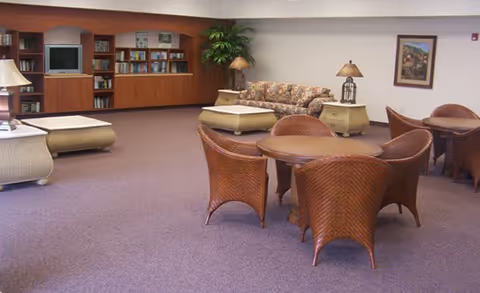 A spacious common area with carpeted floor featuring a round table surrounded by wicker chairs in the foreground. In the background, there is a floral-patterned sofa, two side tables with lamps, a tall plant, and a built-in wooden bookshelf with a television. The walls are light-colored and decorated with a framed picture.