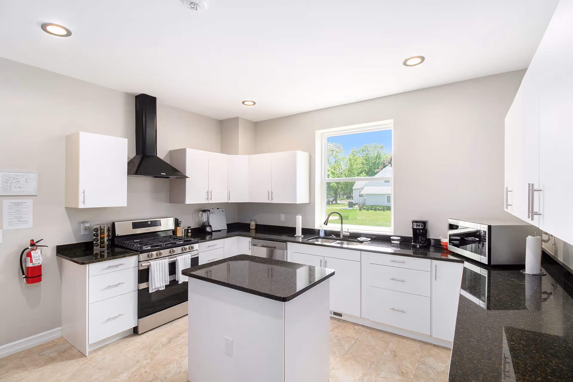 Bright modern kitchen with white cabinets, black countertops, a center island, stainless steel stove and a window overlooking a yard.