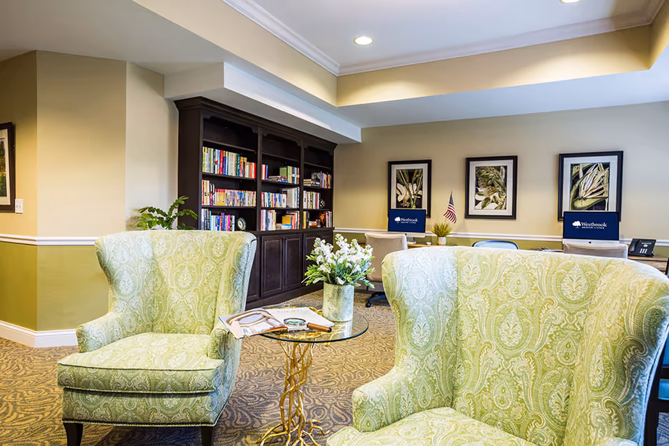 A cozy senior living common area with two green patterned armchairs facing a small glass-top table with a flower vase and reading materials. In the background, there is a dark wooden bookshelf filled with books, a desk with two computer monitors displaying the Westbrook Senior Living logo, and three framed botanical prints on the wall.