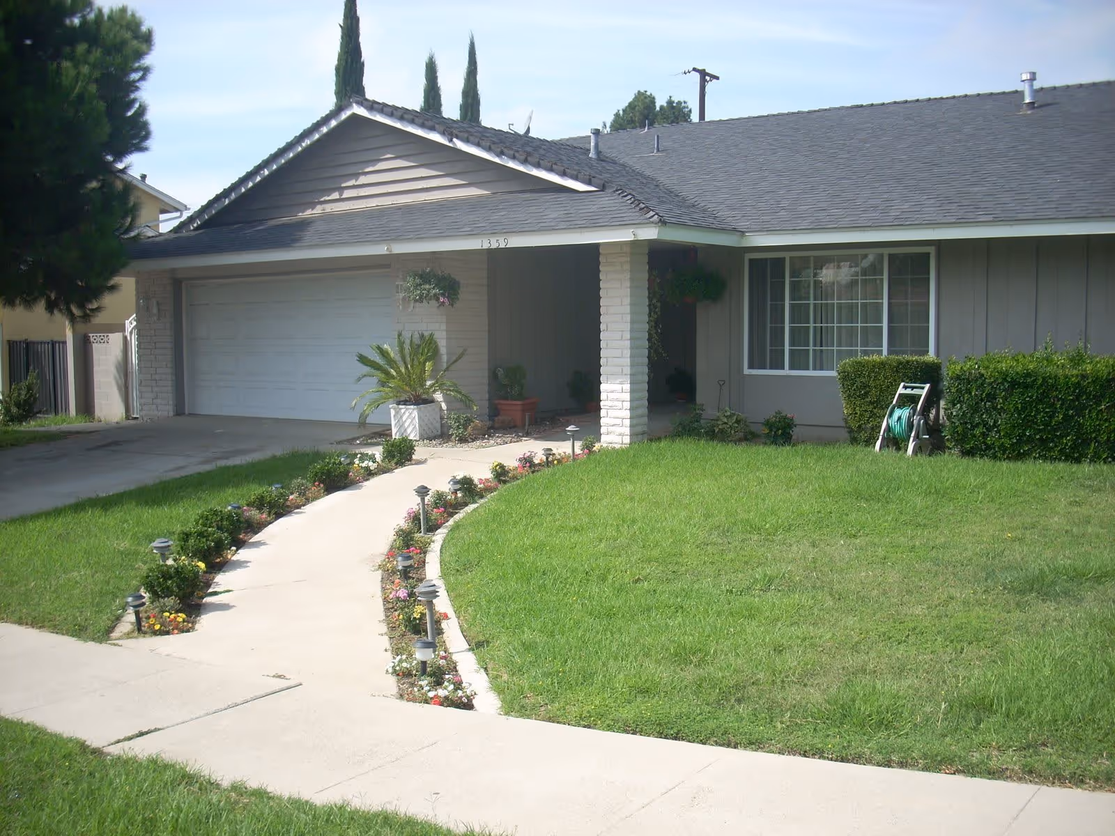 Front exterior view of a single-story residential building with a gray roof, a garage door, a curved concrete walkway lined with small plants and solar lights, a green lawn, and some bushes. The house number 1359 is visible above the garage.