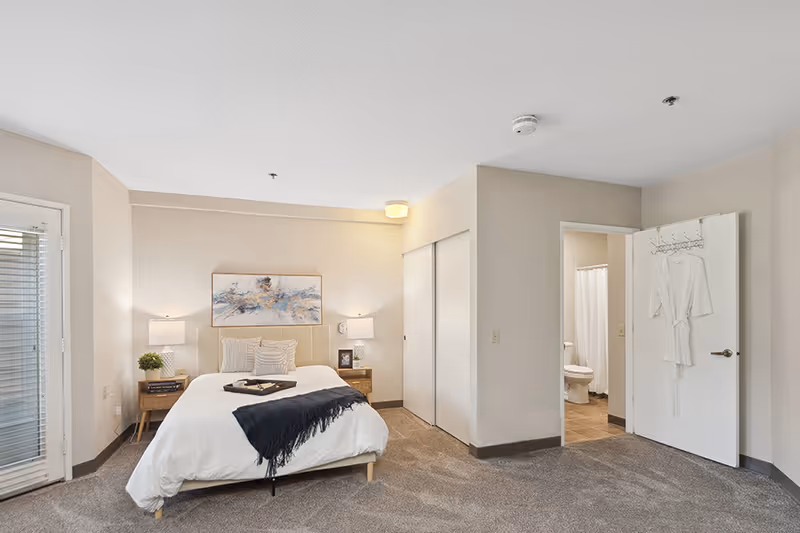 A bright and spacious bedroom with a neatly made bed featuring white bedding and a dark throw blanket. There are two wooden nightstands with lamps and decorative items on either side of the bed. A large abstract painting hangs above the bed. The room has beige walls and carpeted flooring. To the right, there is an open door leading to a bathroom with a visible toilet and shower curtain. Another door has a white robe hanging on a hook. A glass door with blinds is on the left side of the room.