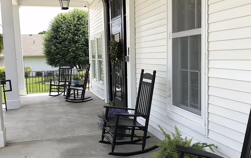 A covered outdoor porch area with black rocking chairs placed along the white exterior wall of a building. There are potted plants near the chairs and a black door with a wreath. In the background, a green lawn and a tree are visible.