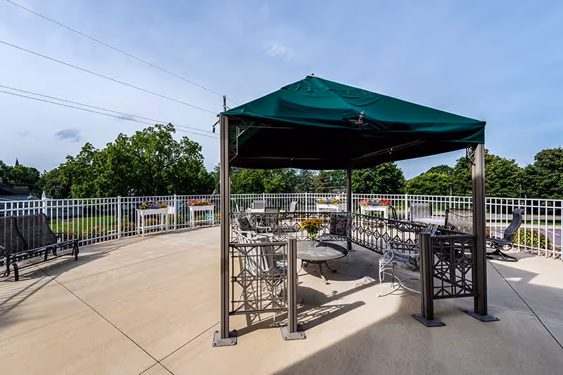 Outdoor patio area with a green canopy covering metal chairs and tables. The patio is surrounded by a white metal railing with flower boxes attached. Trees and a clear blue sky are visible in the background.