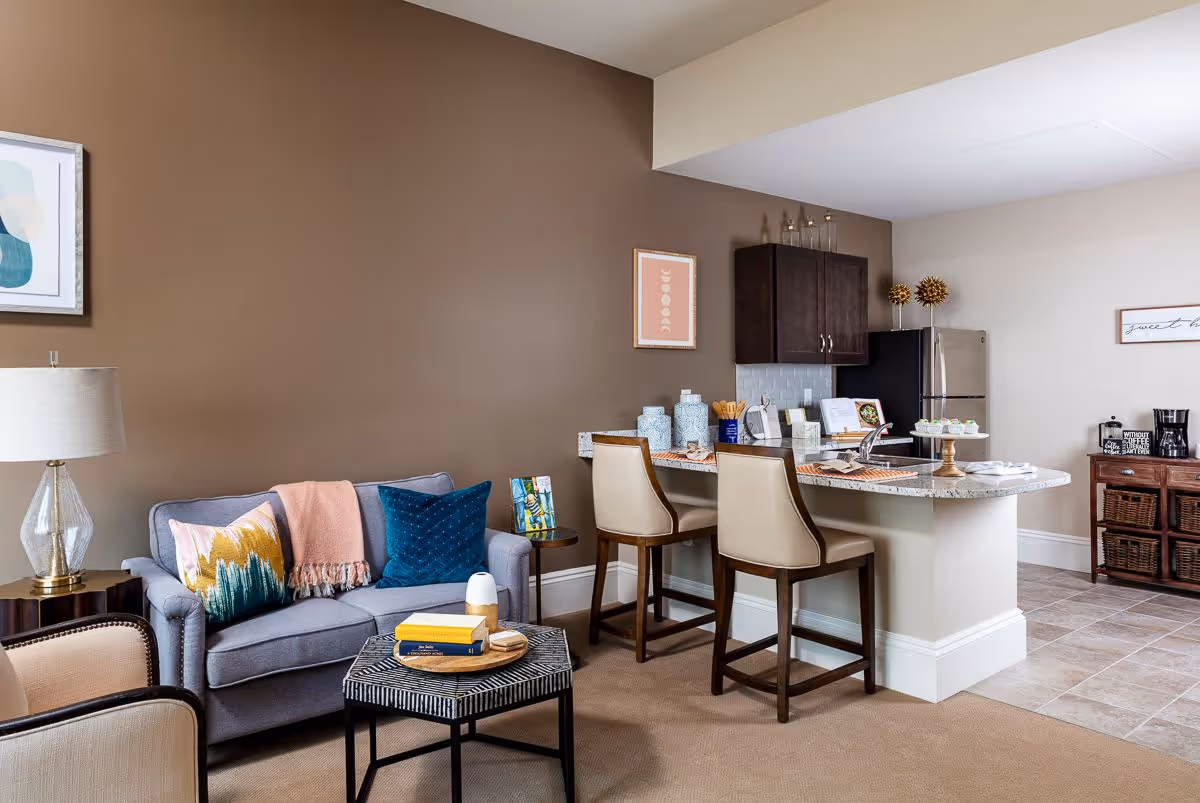 A cozy living area with a gray sofa adorned with colorful pillows and a pink throw blanket. A small round side table with a lamp is beside the sofa. In front, there is a hexagonal coffee table with books and a small decorative item. Adjacent to the living area is a kitchen counter with two beige cushioned bar stools. The kitchen features dark wood cabinets, a stainless steel refrigerator, and various decorative items on the counter. The floor transitions from carpet in the living area to tile in the kitchen.