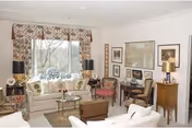 A bright and cozy living room with a large window covered by floral curtains. The room features a white sofa, a glass coffee table, two wooden chairs with cushions, a small wooden side table with a lamp, and framed artwork on the walls. The decor is traditional and inviting.
