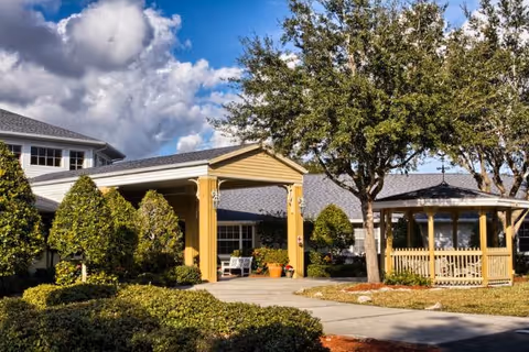 Front entrance of a building with a covered portico, gazebo, trees, and landscaped walkways under a blue sky.