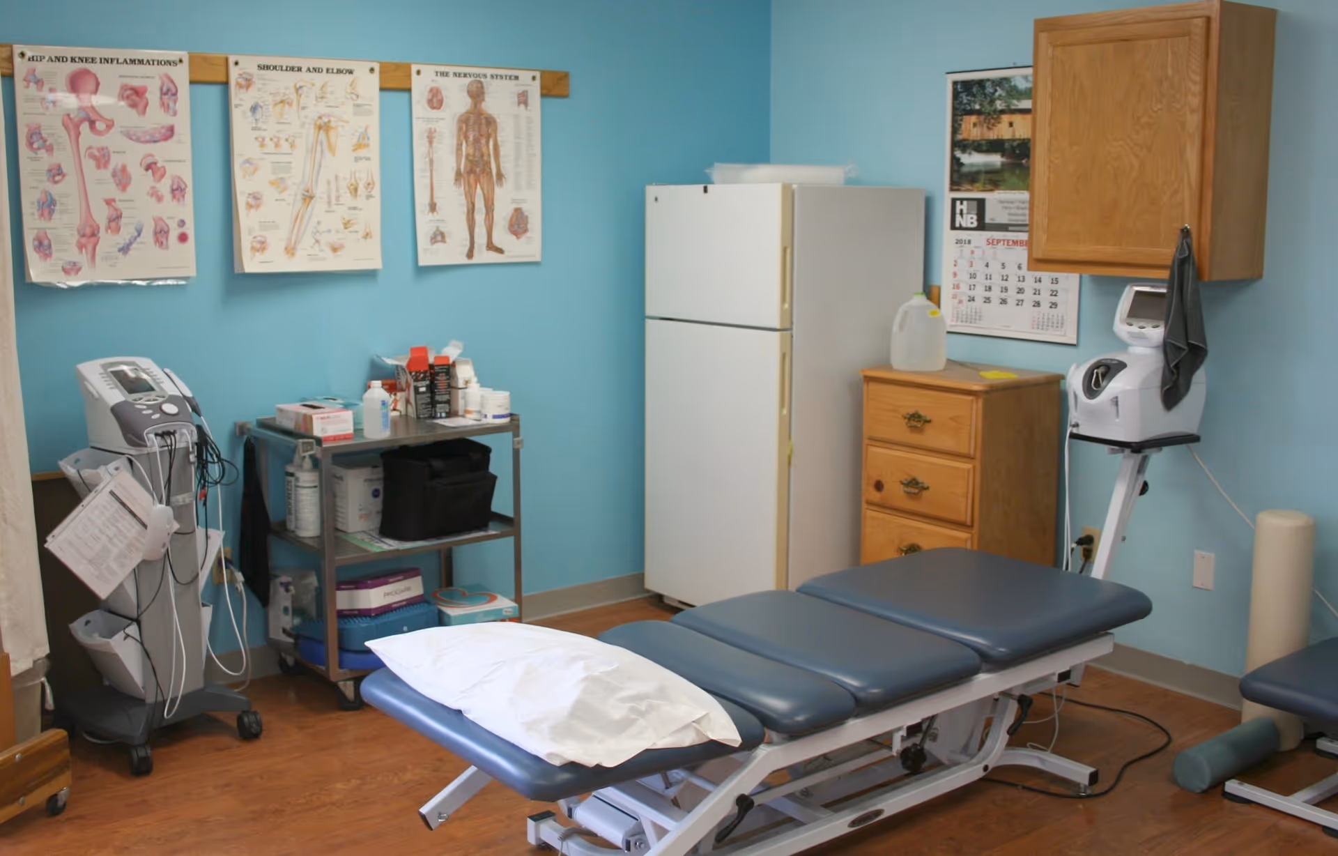 A medical examination or therapy room with a blue padded treatment table, a white pillow, medical equipment on a cart, anatomical charts on a blue wall, a white refrigerator, a wooden cabinet, and a calendar.