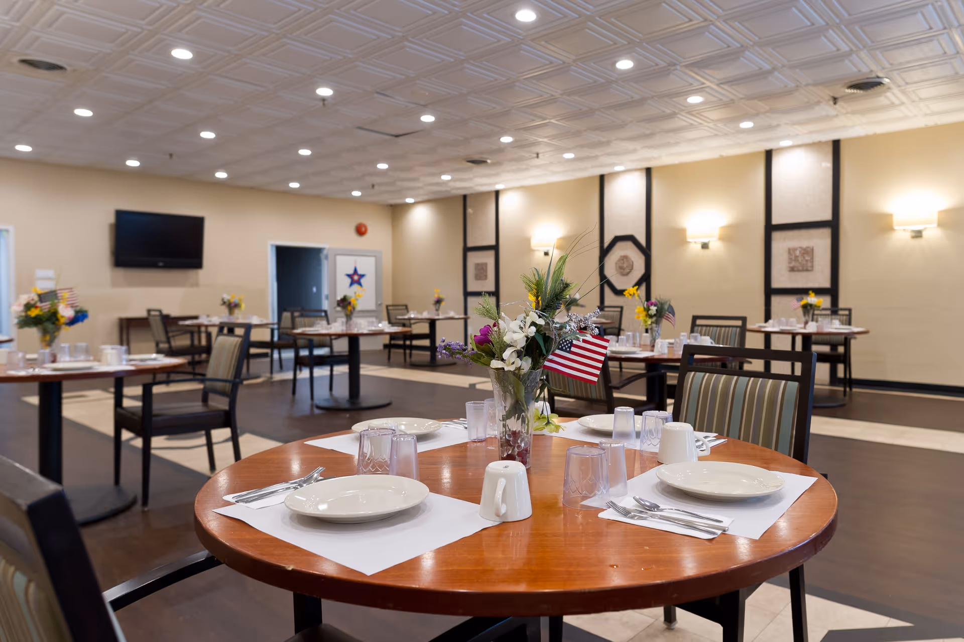 A dining room in a senior living facility with round wooden tables set with white plates, cups, utensils, and placemats. Each table has a vase with a colorful flower arrangement and a small American flag. The room has beige walls, a patterned ceiling with recessed lighting, and a wall-mounted TV. Several chairs with striped cushions are arranged around the tables.