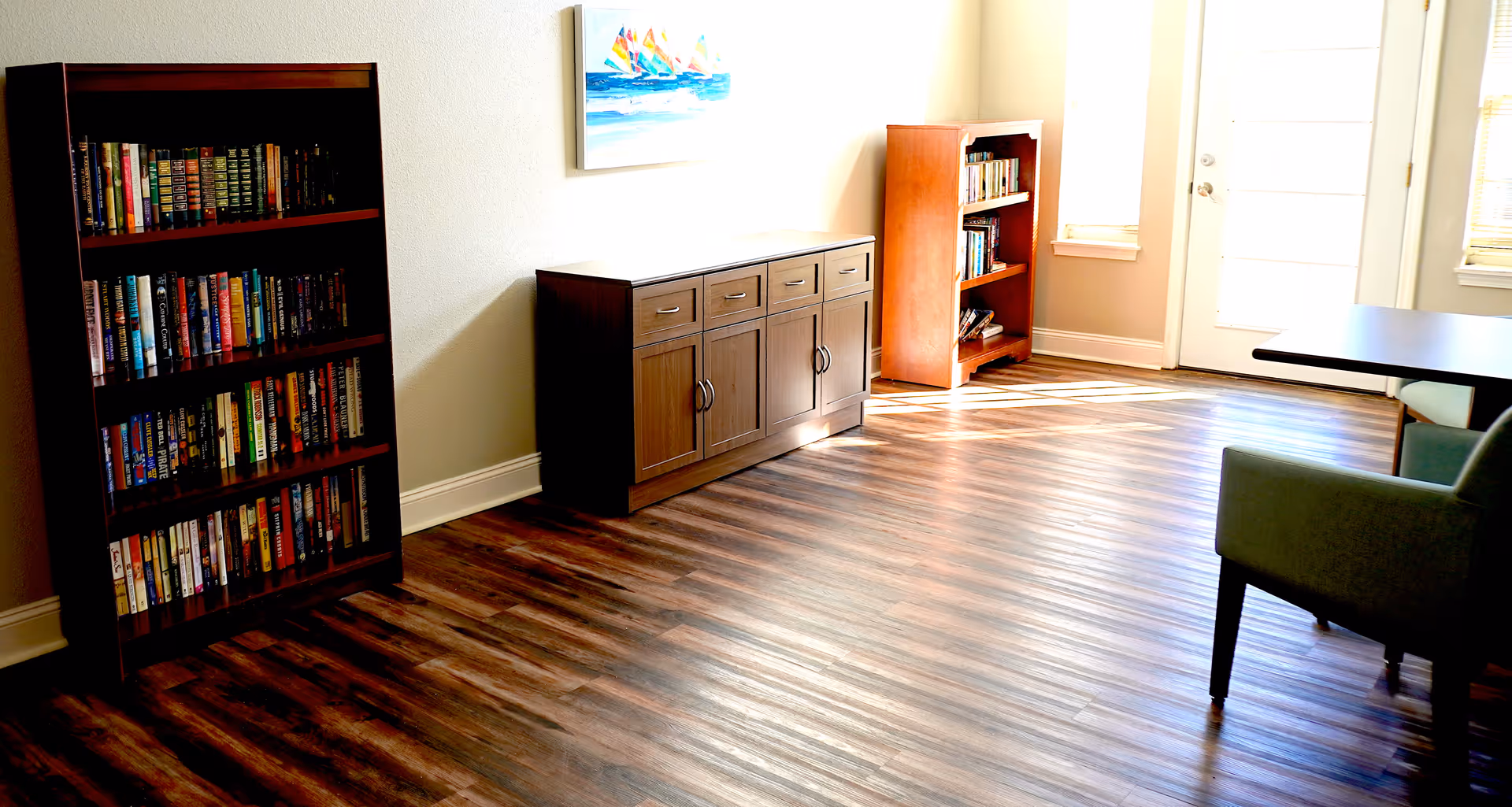 A bright room with wooden flooring featuring two wooden bookshelves filled with books, a wooden cabinet with drawers and doors, a green upholstered chair, a table, and a painting of colorful sailboats on the wall. Sunlight streams in through a glass door and windows.