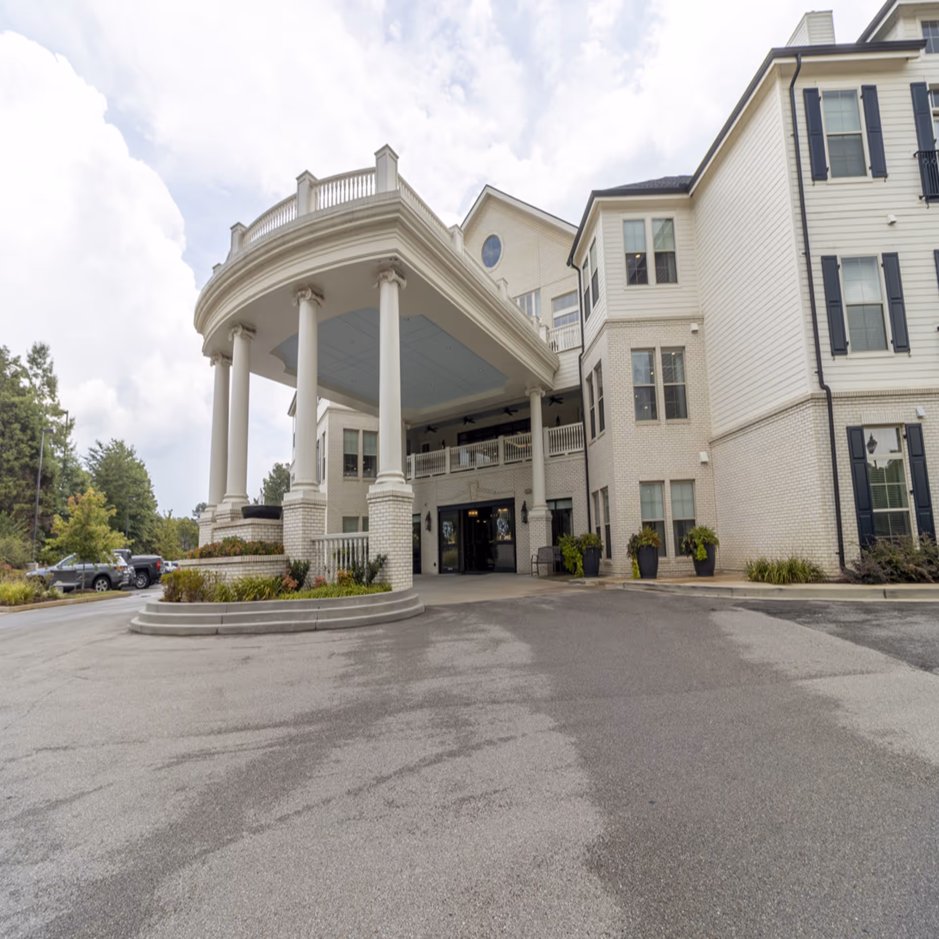Exterior view of a senior living facility entrance with a large covered portico supported by white columns. The building is light-colored with multiple windows and black shutters. There are some plants and parked cars visible on the left side under a cloudy sky.