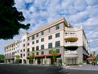 Exterior view of a multi-story senior living facility named The Bellettini with balconies, large windows, and red awnings under a partly cloudy sky.