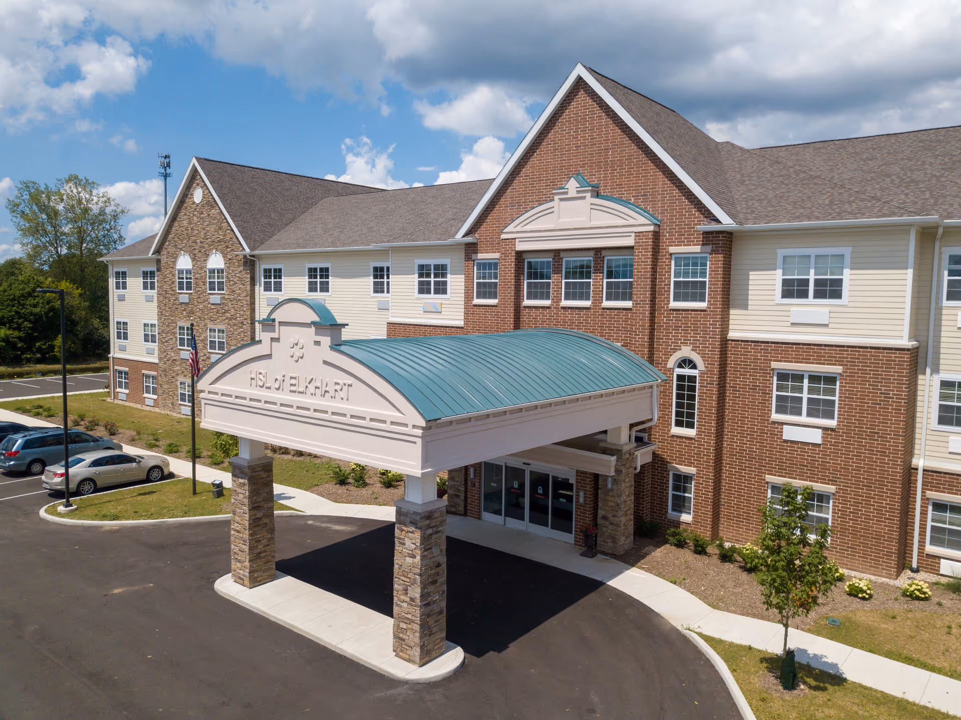 Front entrance and covered porte-cochere of a three-story brick and siding senior living facility under a blue sky.
