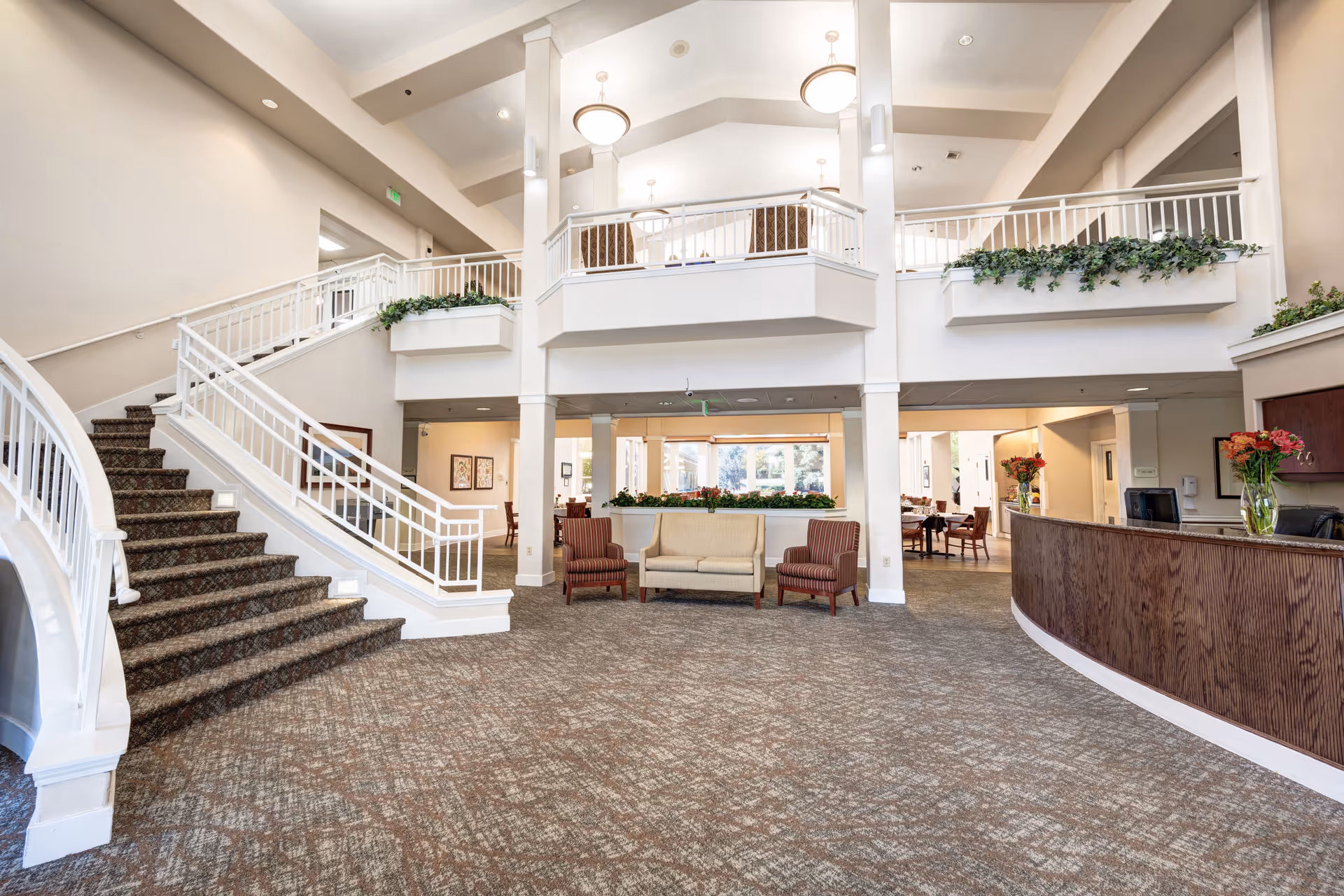 Spacious and well-lit interior lobby area of a senior living facility featuring a carpeted staircase with white railings, a seating area with a beige couch and two striped armchairs, a wooden reception desk with flower vases, and an upper balcony adorned with greenery.