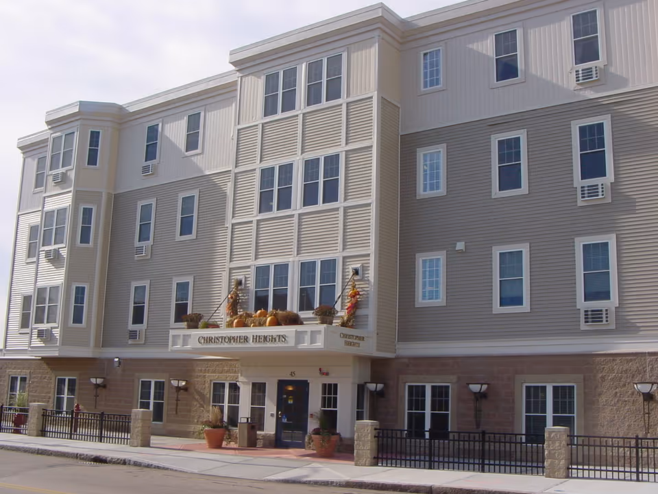 Exterior front view of a multi-story senior living facility named Christopher Heights, featuring beige and light brown siding, multiple windows with air conditioning units, and a decorated entrance with pumpkins and fall-themed decorations.