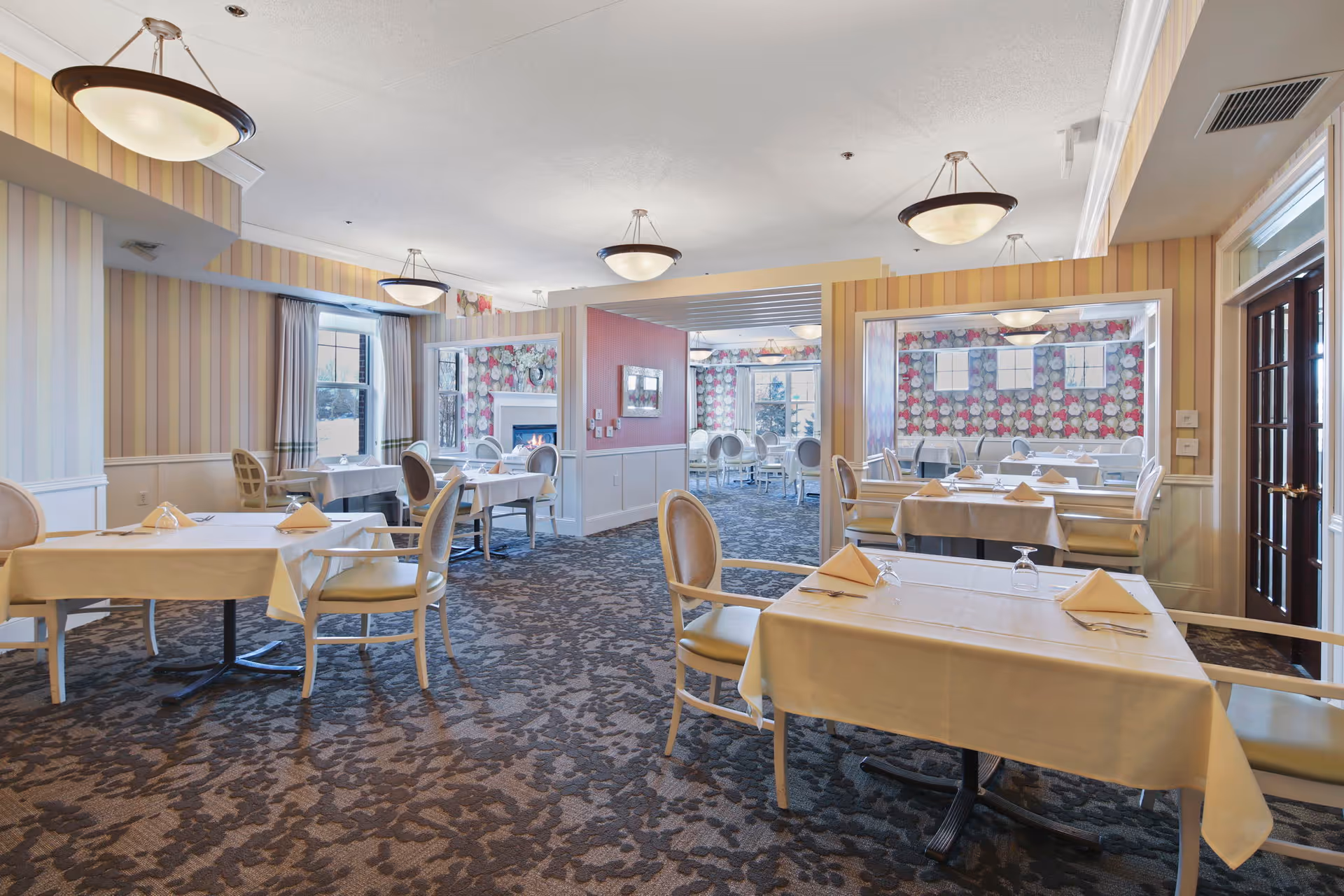 A bright and elegant dining room with several tables covered in white tablecloths, each set with folded napkins and glasses. The room features patterned carpet, striped wallpaper, and large windows with curtains allowing natural light. There are multiple chairs around the tables and decorative light fixtures hanging from the ceiling. In the background, another dining area with floral wallpaper and additional seating is visible.