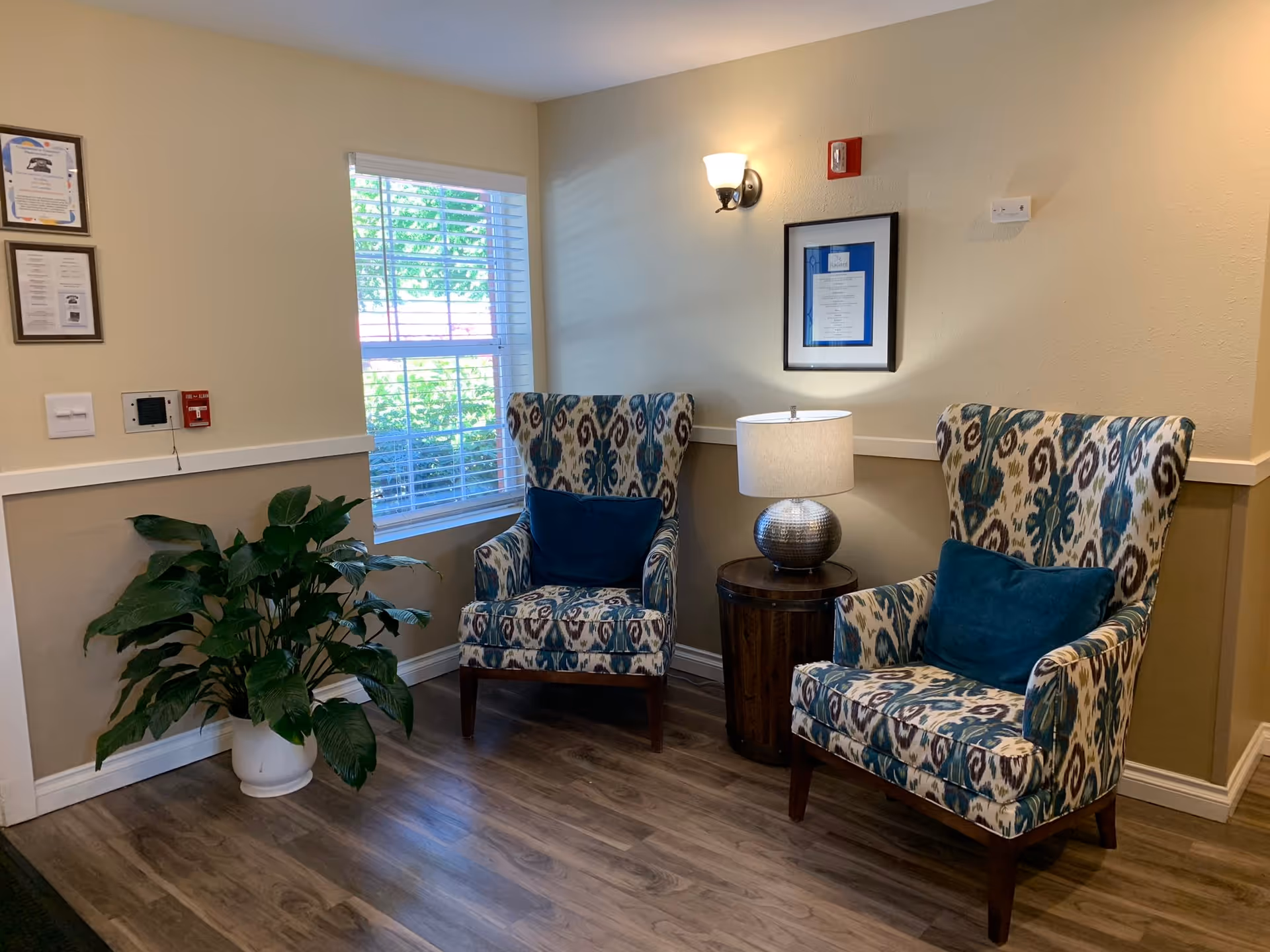 A cozy corner in a senior living facility with two patterned armchairs featuring blue cushions, a round wooden side table with a silver lamp, a large green potted plant, and a window with white blinds letting in natural light. The walls are painted beige with framed certificates and a wall sconce light above.