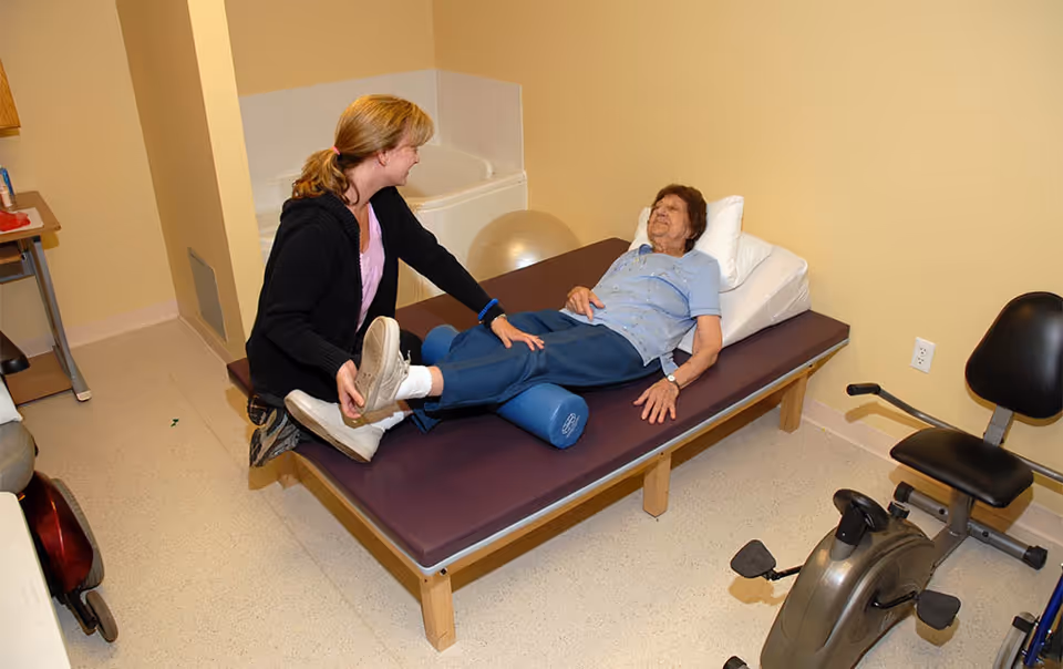 A physical therapist assists an elderly woman lying on a padded therapy table in a rehabilitation room. The therapist is holding and stretching the woman's leg, which is supported by a foam roller. The room has beige walls, a stationary exercise bike, and a large exercise ball in the corner.