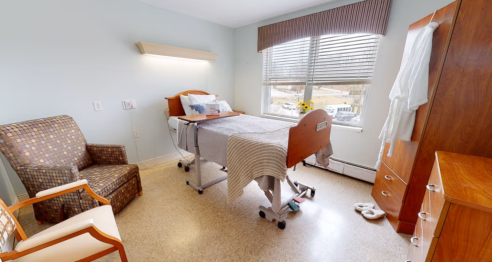 A bright and clean patient room in a healthcare facility featuring a hospital bed with a wooden headboard and footboard, a small overbed table, a patterned armchair, a wooden wardrobe with a white robe hanging on the door, and a window with blinds letting in natural light.