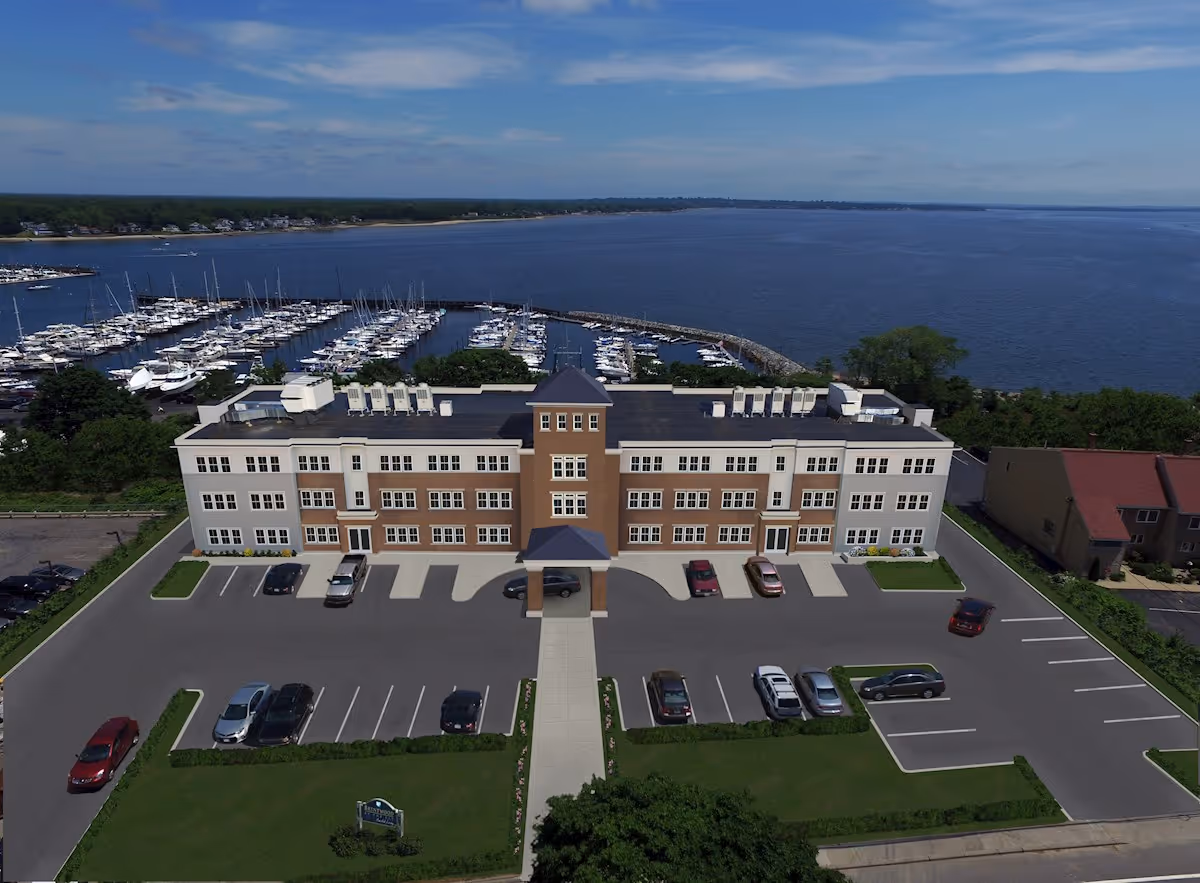 Aerial view of a three-story assisted living facility named Brentwood by the Bay Assisted Living, with a parking lot in front and a marina with numerous boats docked behind the building, adjacent to a large body of water under a partly cloudy sky.
