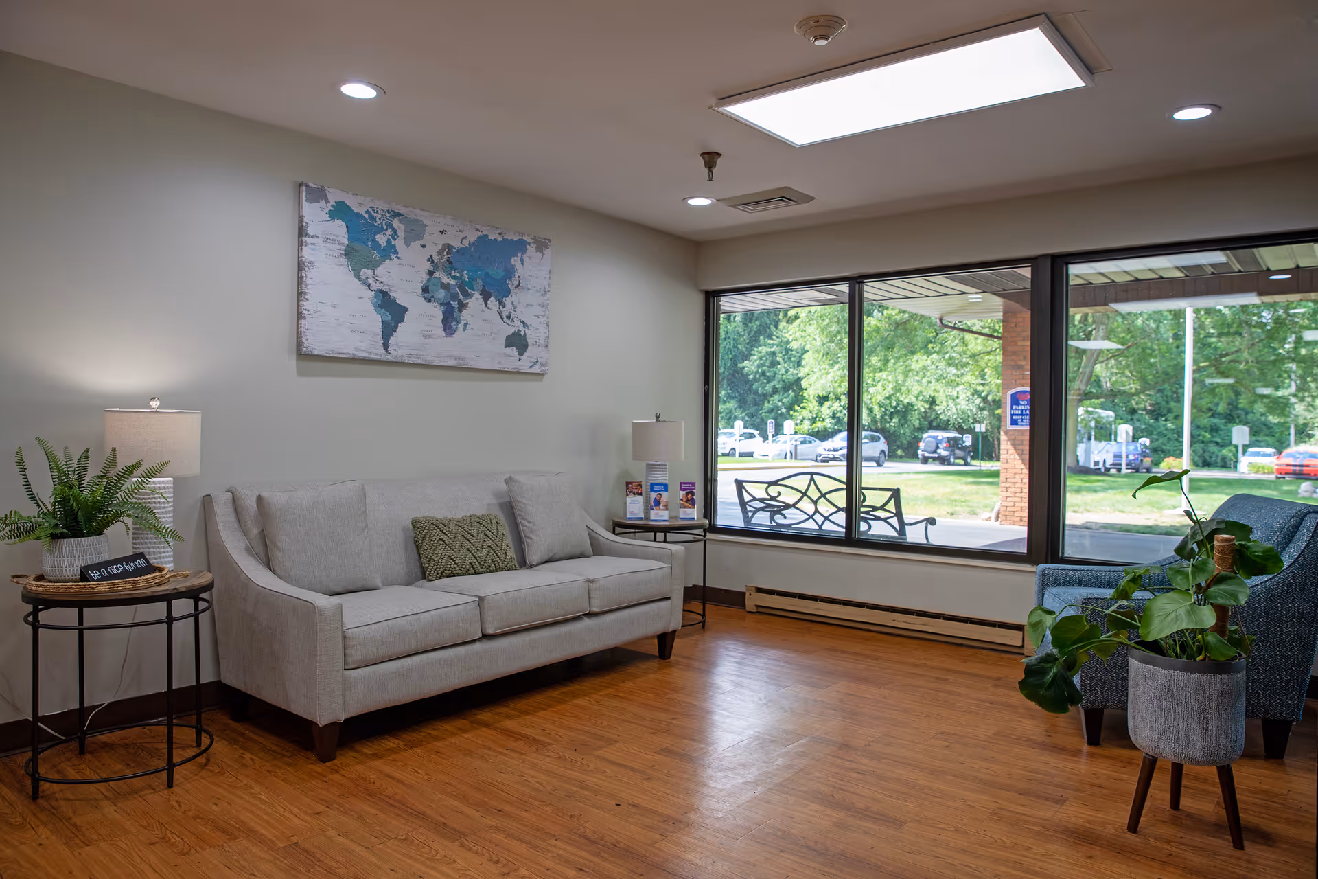 A bright and cozy living room area with a light gray sofa adorned with cushions, a side table with a lamp and a small plant, a large window showing an outdoor view with trees and parked cars, a blue armchair, and a potted plant on the wooden floor. A world map artwork hangs on the wall above the sofa.
