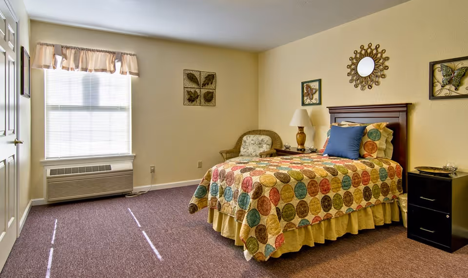 A cozy bedroom with a single bed covered in a colorful circular patterned quilt and a blue pillow. There is a wooden headboard, a nightstand with a lamp, and a wicker chair with a floral cushion. The walls are decorated with framed artwork and a decorative mirror. A window with blinds and a valance lets in natural light, and there is a heating/cooling unit below the window.