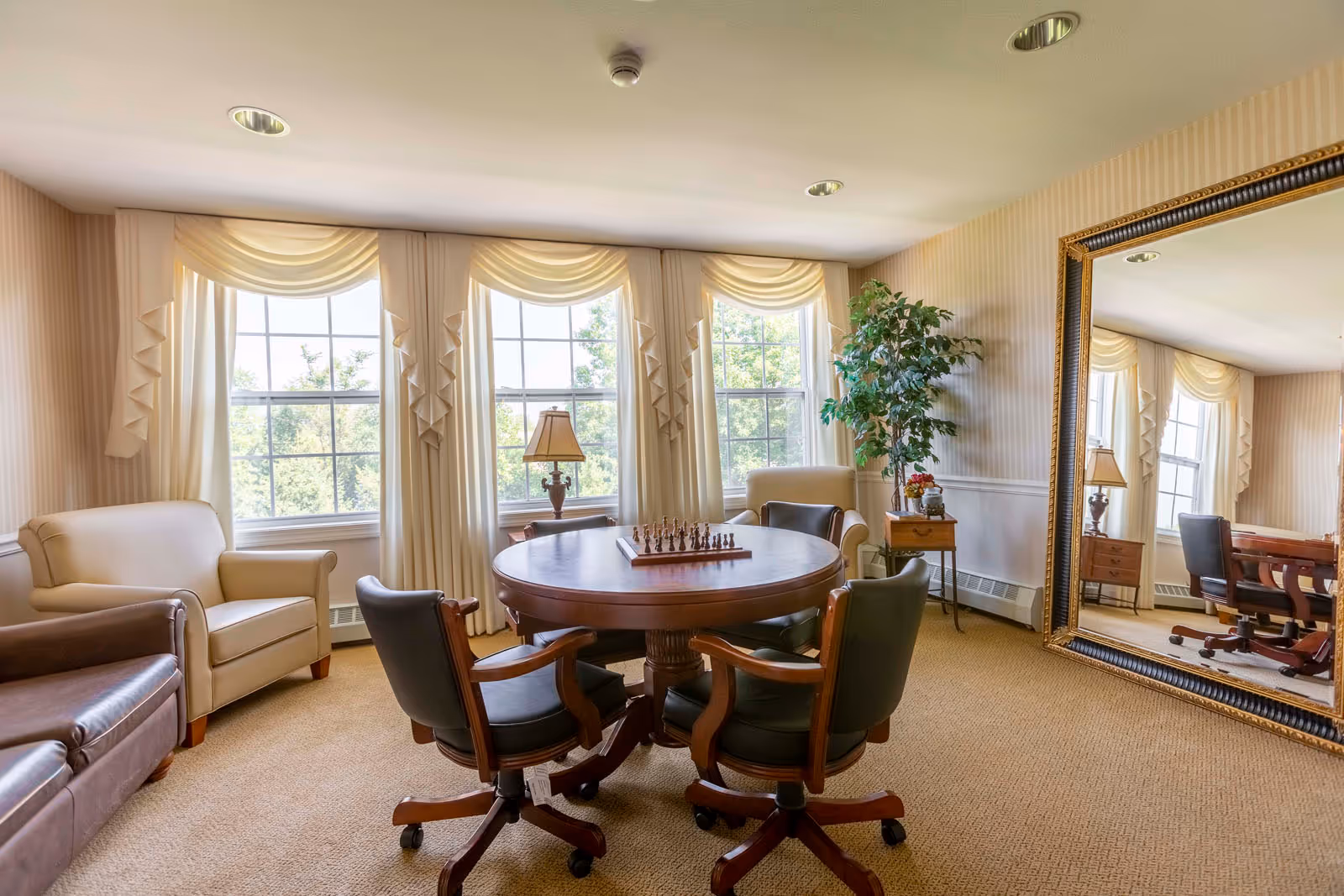 A cozy sitting room with a round wooden table in the center featuring a chess set. Four cushioned chairs with wooden arms surround the table. The room has large windows with cream-colored drapes allowing natural light to fill the space. There are two armchairs, one beige and one brown, placed near the windows. A large ornate mirror is mounted on the right wall, reflecting part of the room. A small side table with a lamp and a potted plant add to the warm and inviting atmosphere.