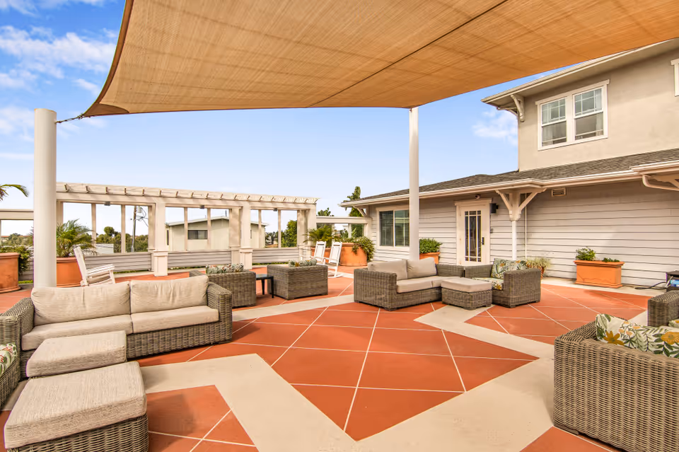 Outdoor patio area with cushioned wicker sofas and chairs arranged on a red and beige tiled floor under a large beige shade sail. The patio is adjacent to a light gray building with white trim and large windows, and there are large potted plants around the seating area.