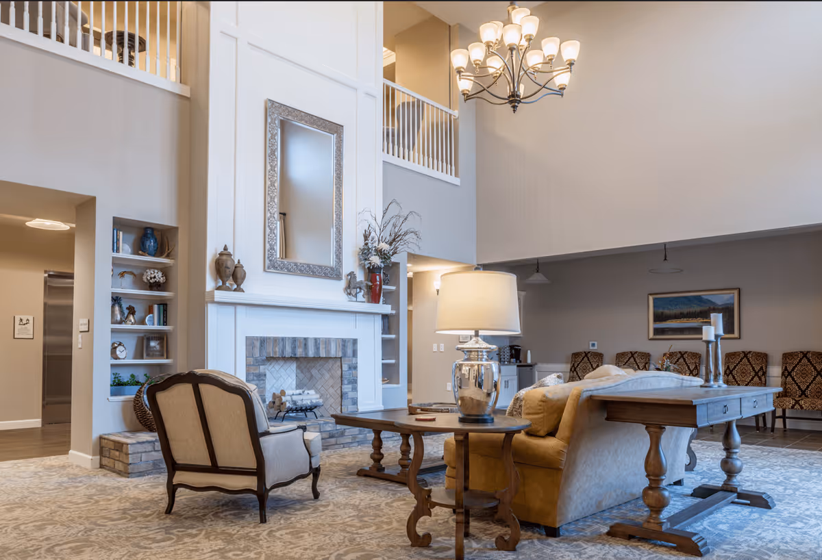 A spacious and well-lit senior living facility common area with a high ceiling, a large fireplace with a decorative mirror above it, comfortable seating including an armchair and a sofa, a wooden coffee table, a side table with a lamp, and a row of patterned chairs against the far wall. The room features neutral tones and elegant decor.
